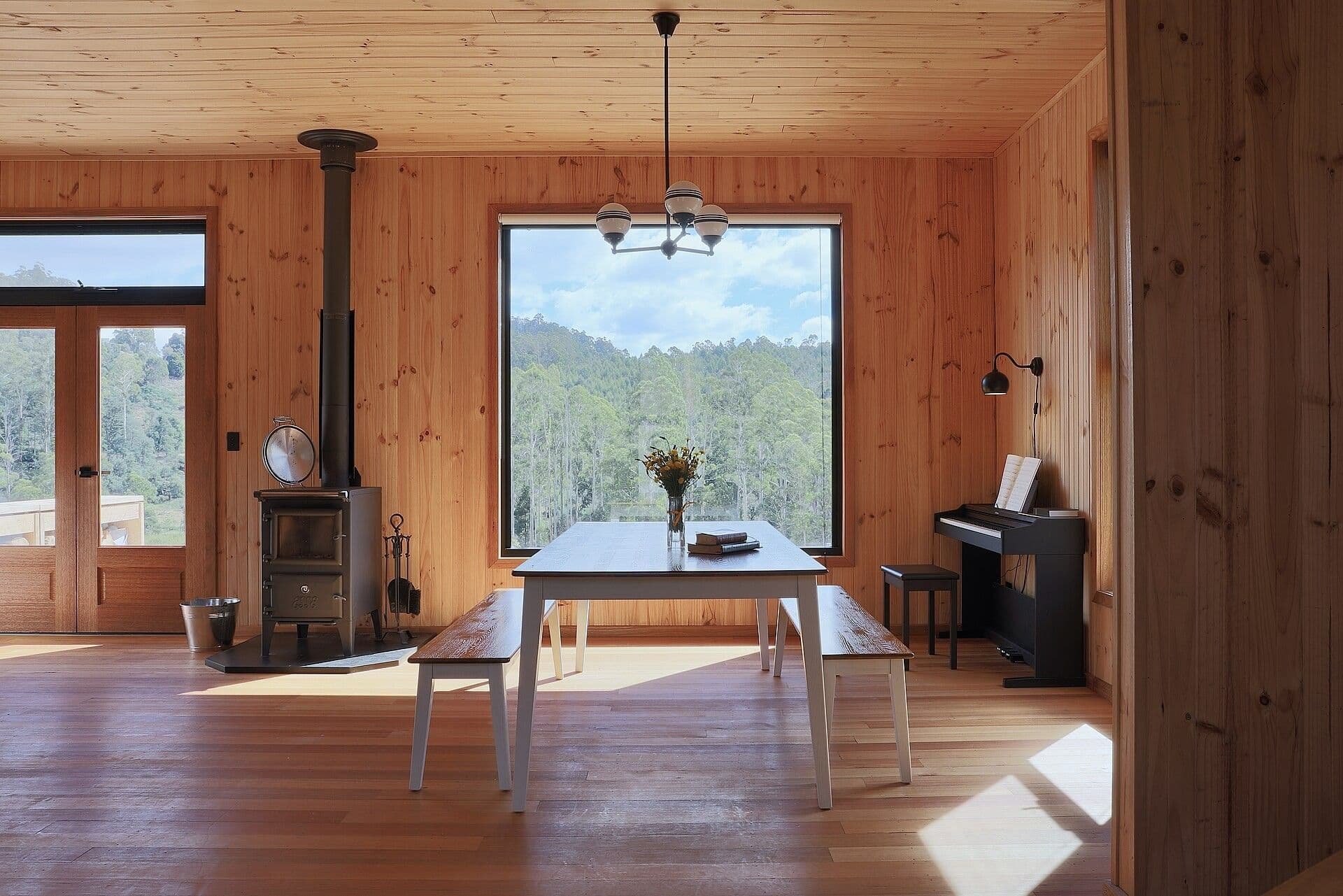 A timber lined dining room with a wood heater and a large window onto trees.